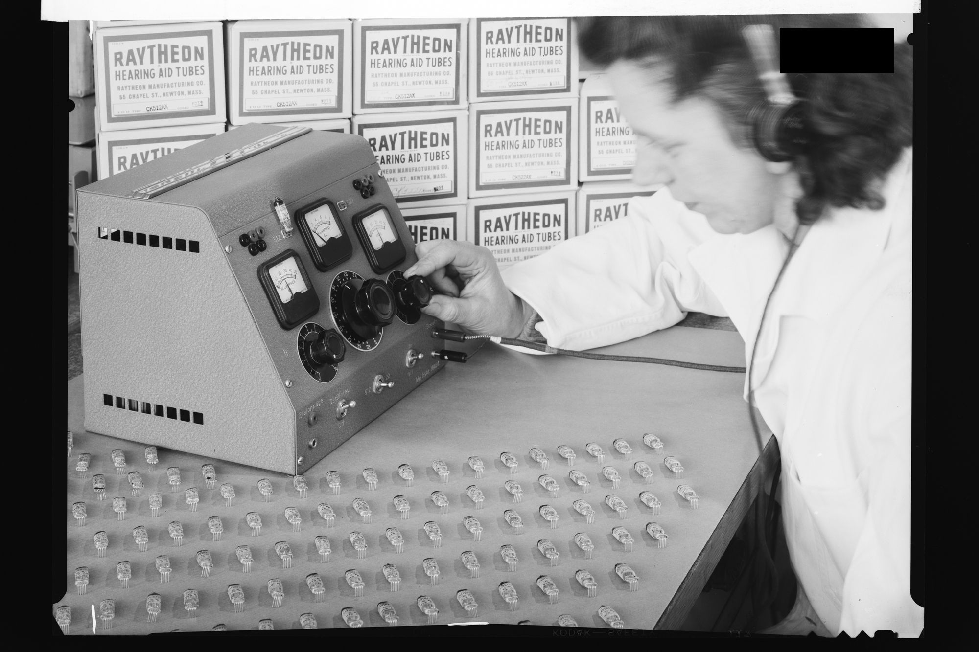 Schwarzweiss-Fotografie. Eine Frau mit Kopfhörer prüft an einem Messgerät Röhren, dahinter Schachteln mit der Aufschrift Raytheon Hearing Aid Tubes.