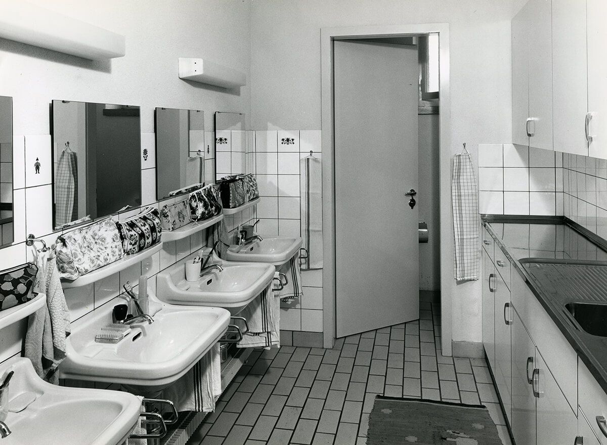 Schwarzweiss-Fotografie des gekachelten Waschraums im Pestalozziheim Redlikon, um 1964. Eine Reihe Waschbecken mit Spiegeln, darüber hängen Handtücher.