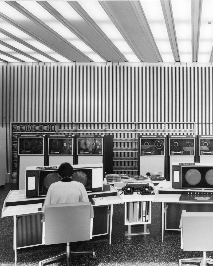 Schwarzweiss-Fotografie eines Computerraums von 1970. Eine Frau sitzt an einem Terminal, dahinter stehen Magnetbandgeräte in einer Reihe.